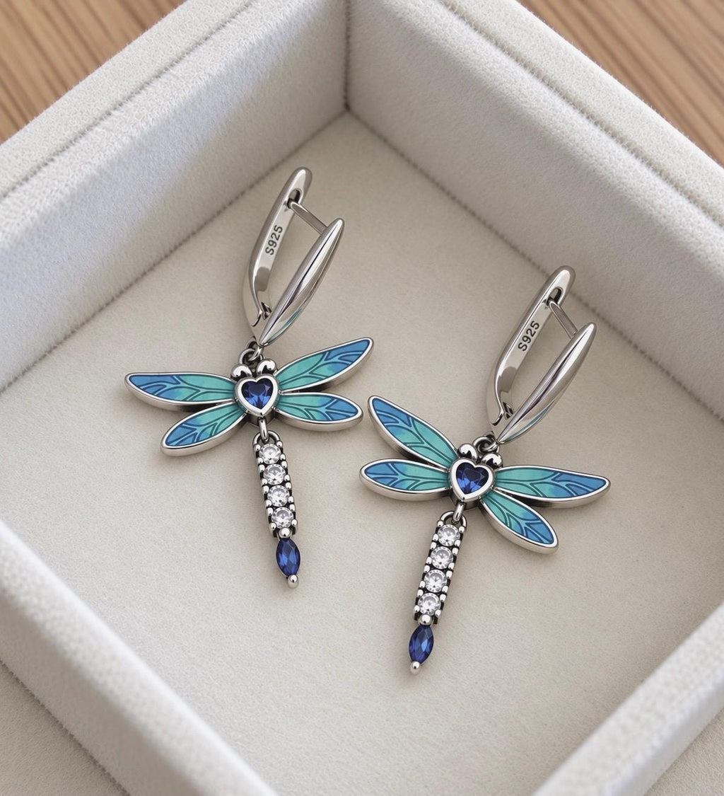 Pair of 925 sterling silver blue enamel dragonfly drop earrings with heart-shaped blue centre stones and sparkling accents on ivory jewellery display