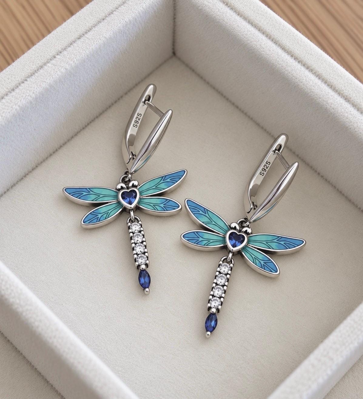Pair of 925 sterling silver blue enamel dragonfly drop earrings with heart-shaped blue centre stones and sparkling accents on ivory jewellery display