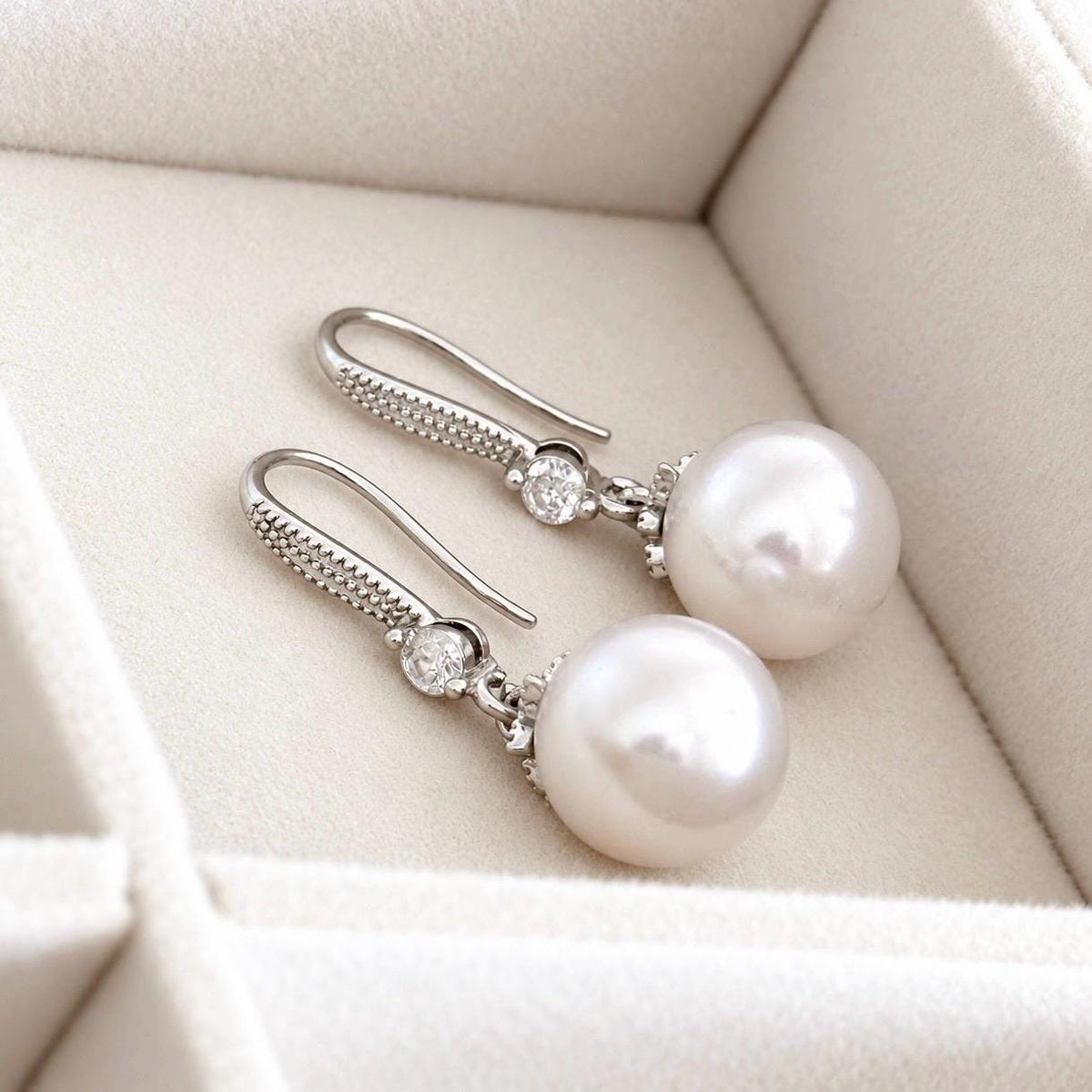 Pair of pearl drop hook earrings lying on cream velvet showing silver hooks and clear crystal accents