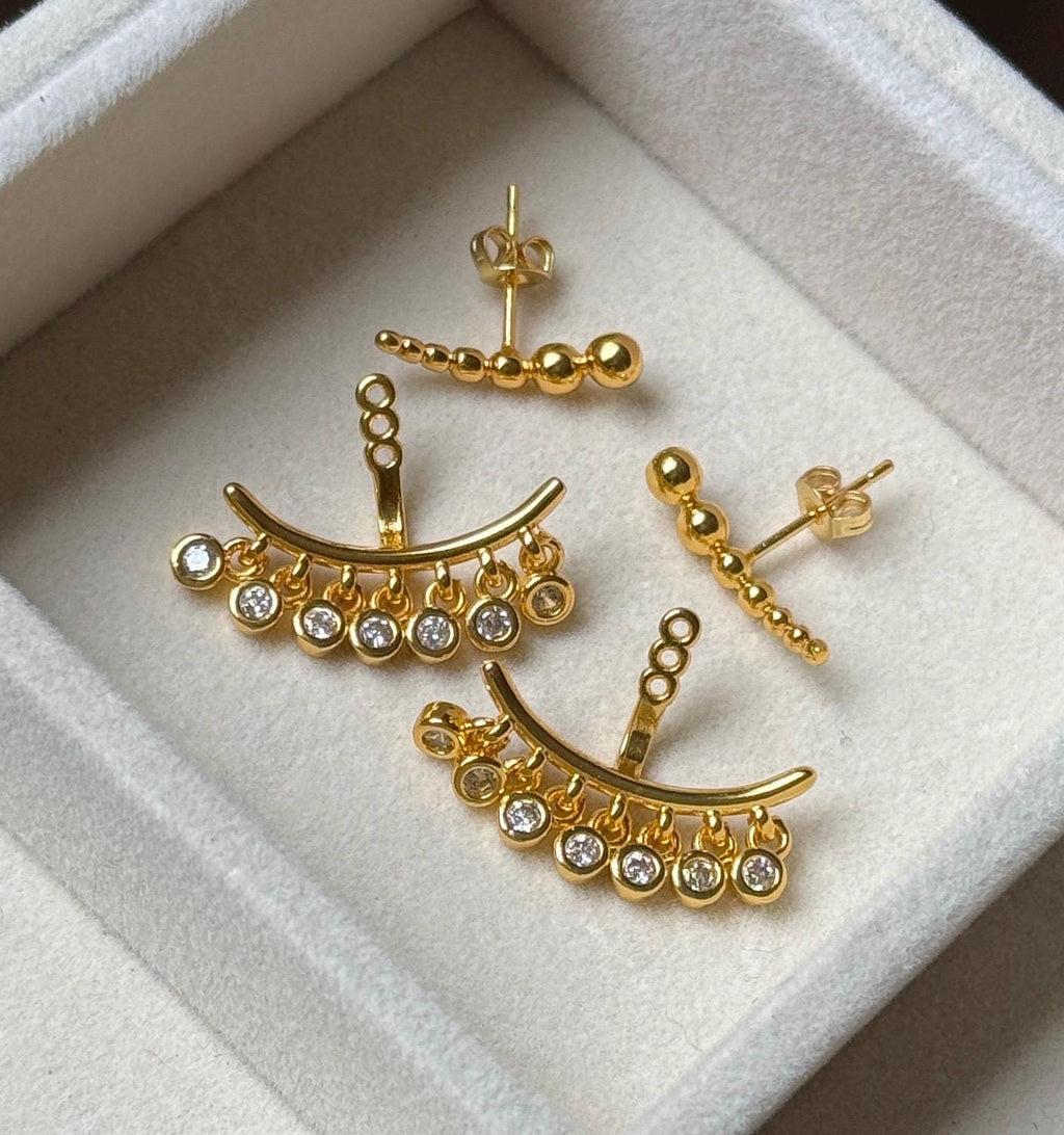 Pair of gold tone stainless steel ear jacket earrings with ball stud fronts and CZ dangling jacket backs displayed on cream velvet