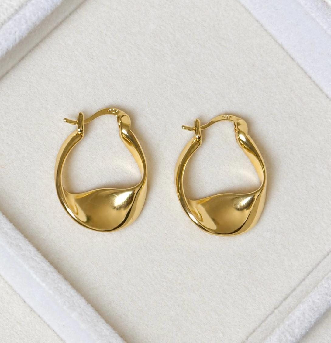 Matching pair of polished gold plated organic teardrop hoop earrings laid on cream velvet