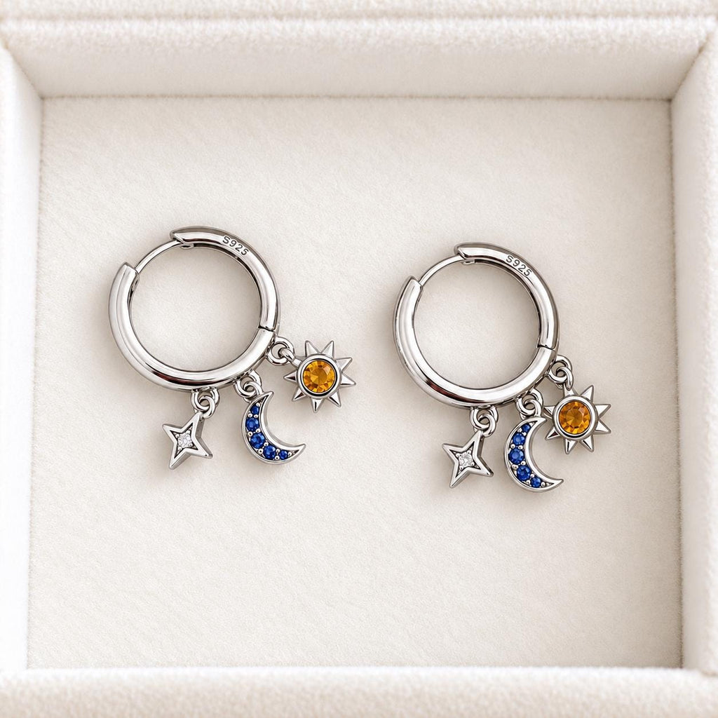 Pair of sterling silver celestial huggie earrings with dangling star, blue crescent moon and amber sun charms on white velvet