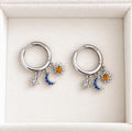 Pair of sterling silver celestial huggie earrings with dangling star, blue crescent moon and amber sun charms on white velvet