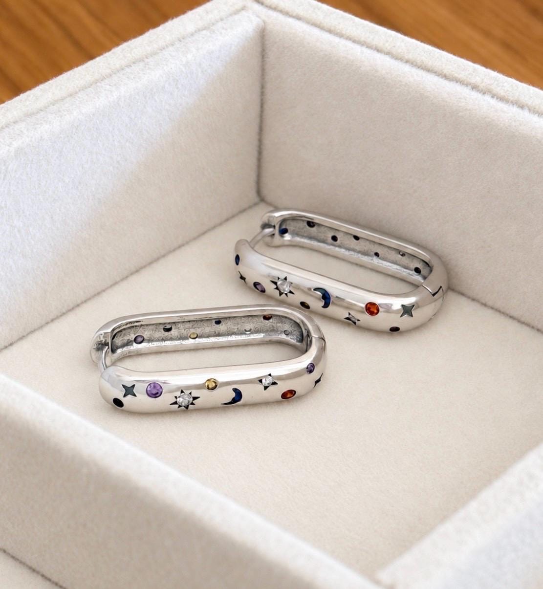 Pair of 925 sterling silver celestial oval hoops shown at an angle in cream jewellery display