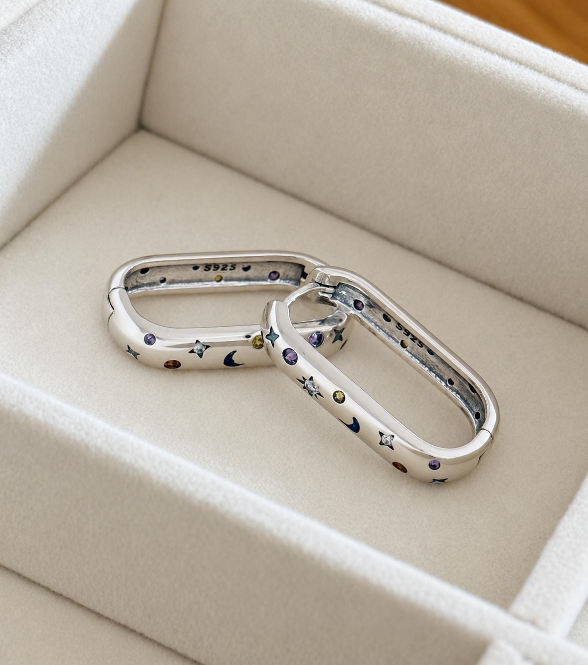 Close-up of silver elongated oval celestial hoops with multicoloured stones, moon and star decorations and S925 marking
