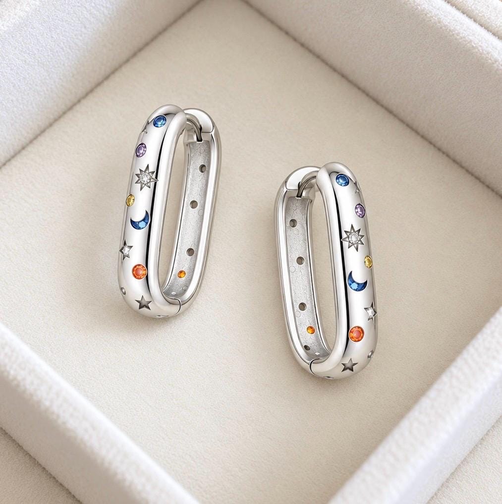 Pair of polished 925 sterling silver elongated hoop earrings with colourful CZ accents, sun moon and star motifs on ivory jewellery background