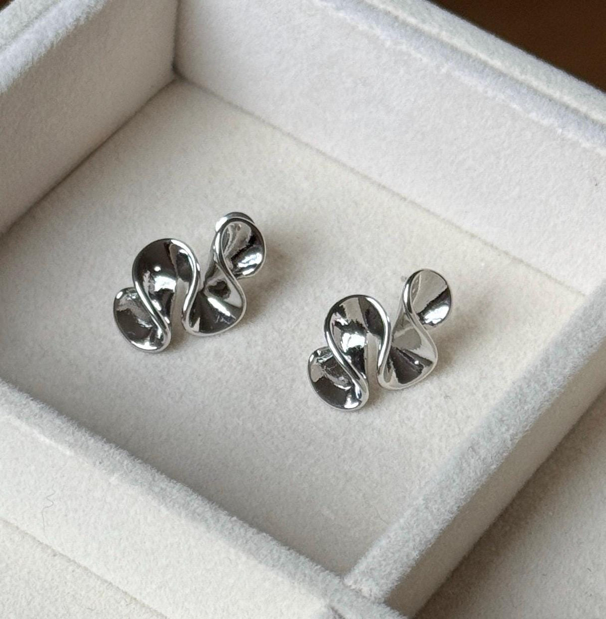 Pair of polished silver-colour folded ribbon sculptural stud earrings on cream velvet