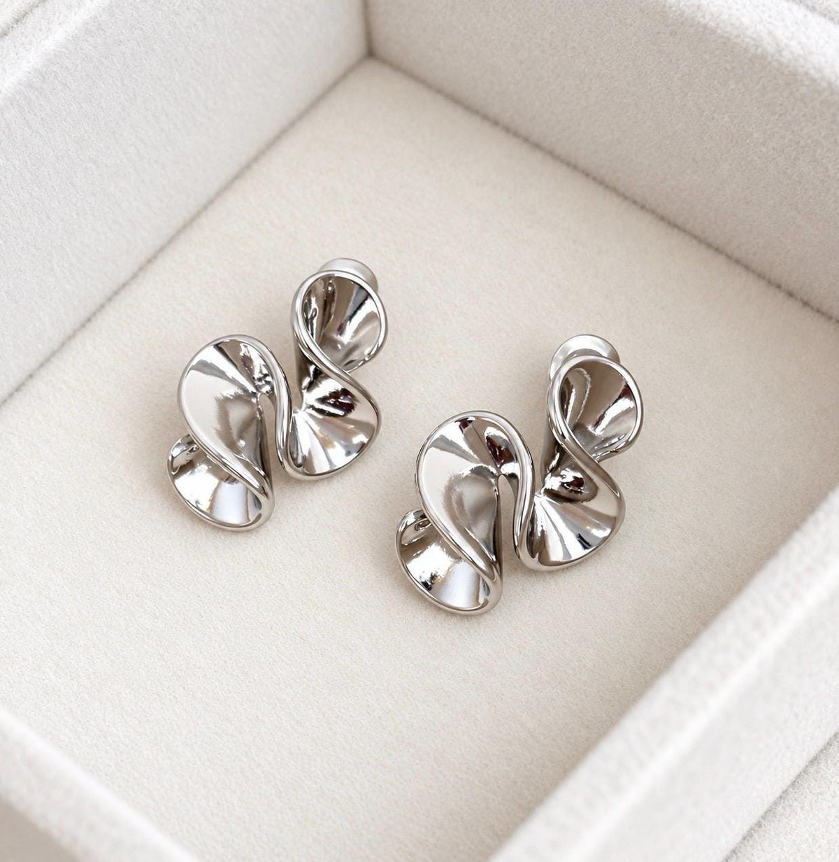 Pair of polished silver-colour abstract sculptural swirl stud earrings on soft cream velvet