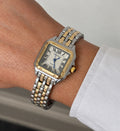 Two-tone gold and silver square Roman numeral watch worn on a wrist with a white sleeve